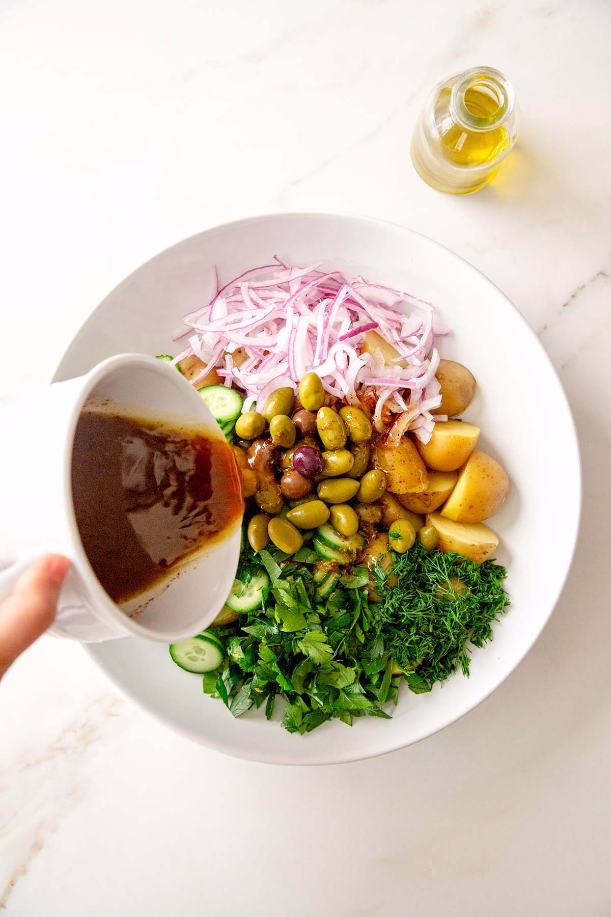 Sumac dressing being poured over chopped potatoes, olives, red onion, cucumber, parsley, and dill in a white mixing bowl