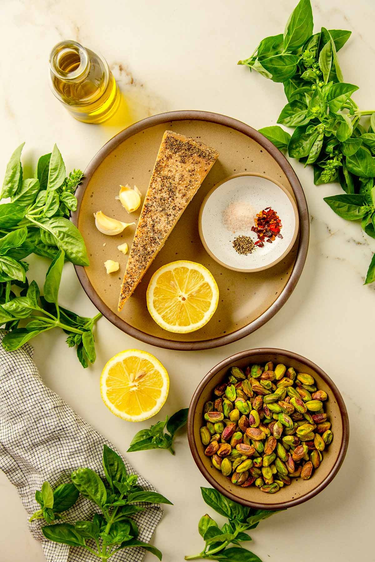 Overhead view of fresh basil, shelled pistachios, Parmesan cheese wedge, garlic cloves, lemon halves, olive oil, and seasonings arranged on a light marble surface.