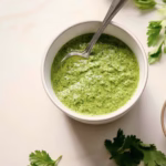 Spicy cilantro sauce (Zhoug) served in a bowl with a spoon, next to fresh garlic and herbs.