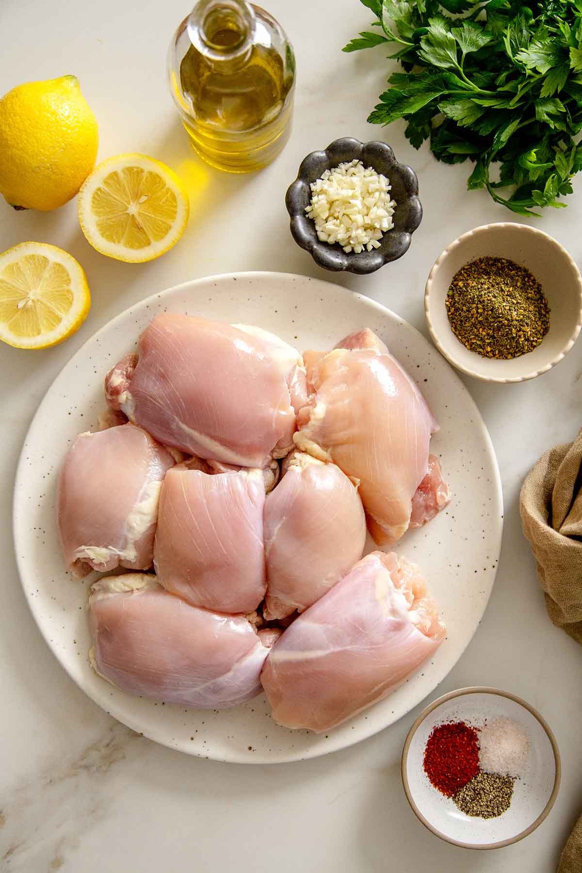 Raw chicken thighs on a plate surrounded by marinade ingredients: lemon, olive oil, garlic, za'atar, and spices.