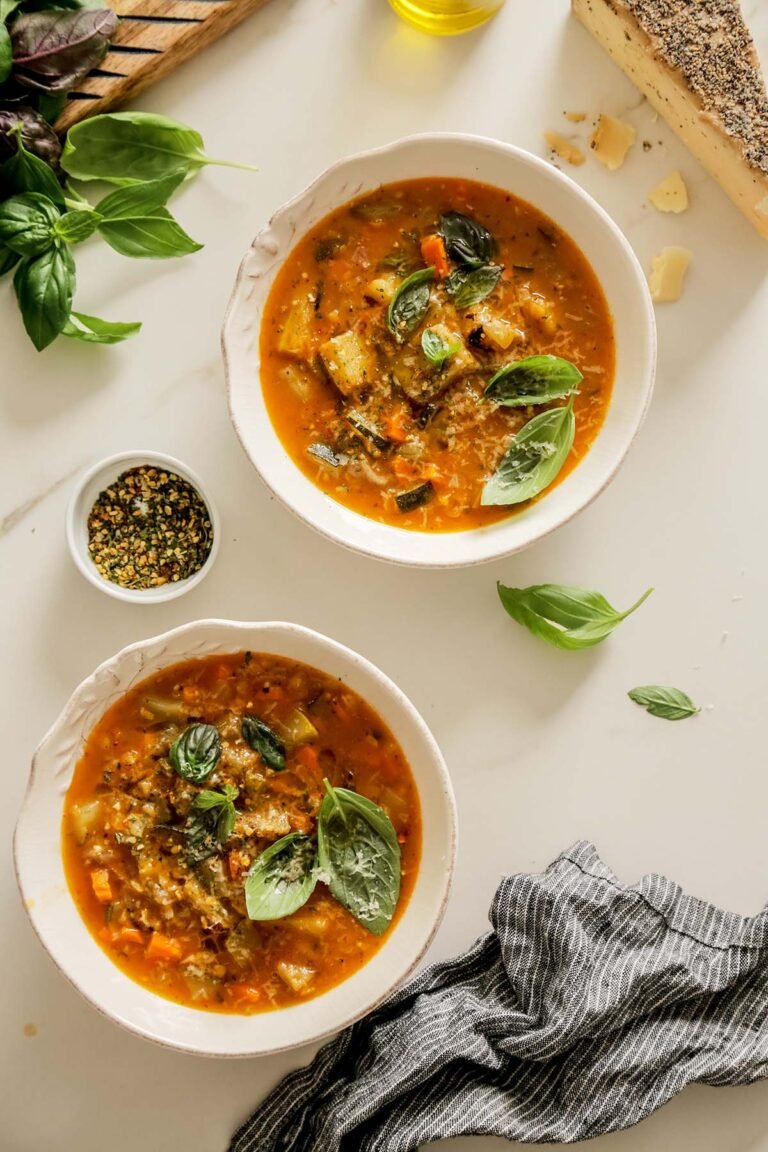 Two servings of rustic roasted vegetable soup in white bowls, garnished with basil and cheese.