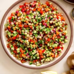 Vibrant Shirazi salad in a white bowl with a brown rim, featuring finely diced cucumbers, tomatoes, red onions, and fresh mint, viewed from an overhead perspective. Lemon wedges are on the side.