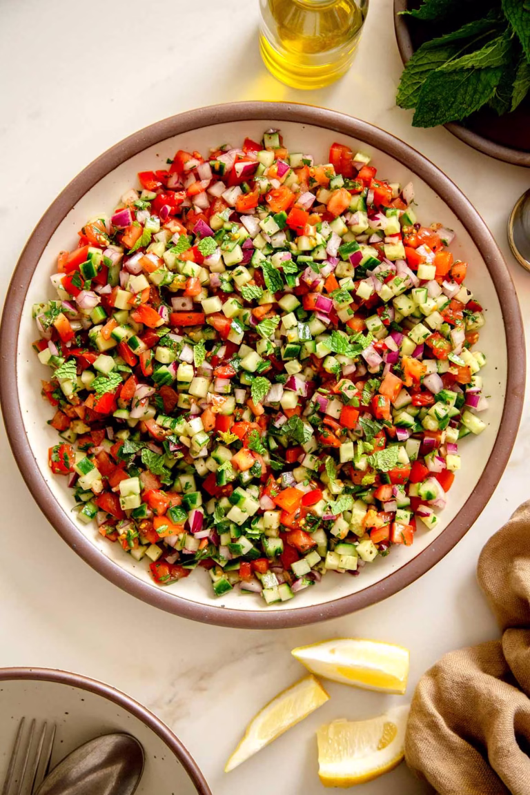 Vibrant Shirazi salad in a white bowl with a brown rim, featuring finely diced cucumbers, tomatoes, red onions, and fresh mint, viewed from an overhead perspective. Lemon wedges are on the side.