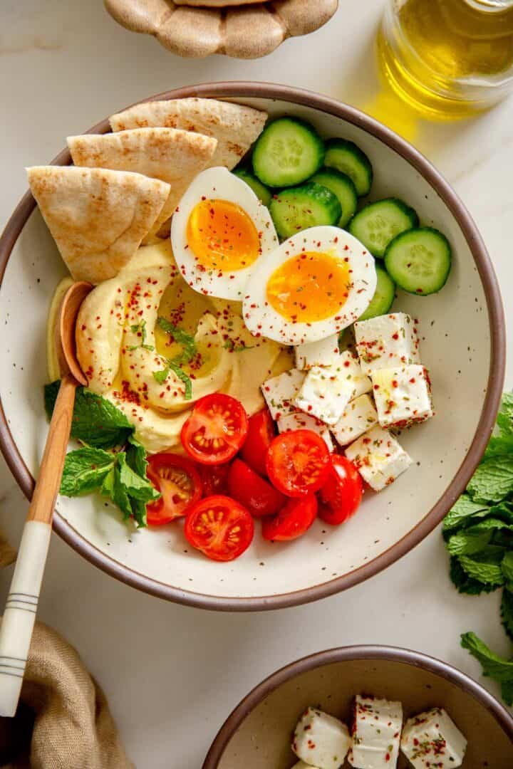 Close-up of a Mediterranean Breakfast Bowl with a spoon, highlighting the creamy hummus and fresh toppings.