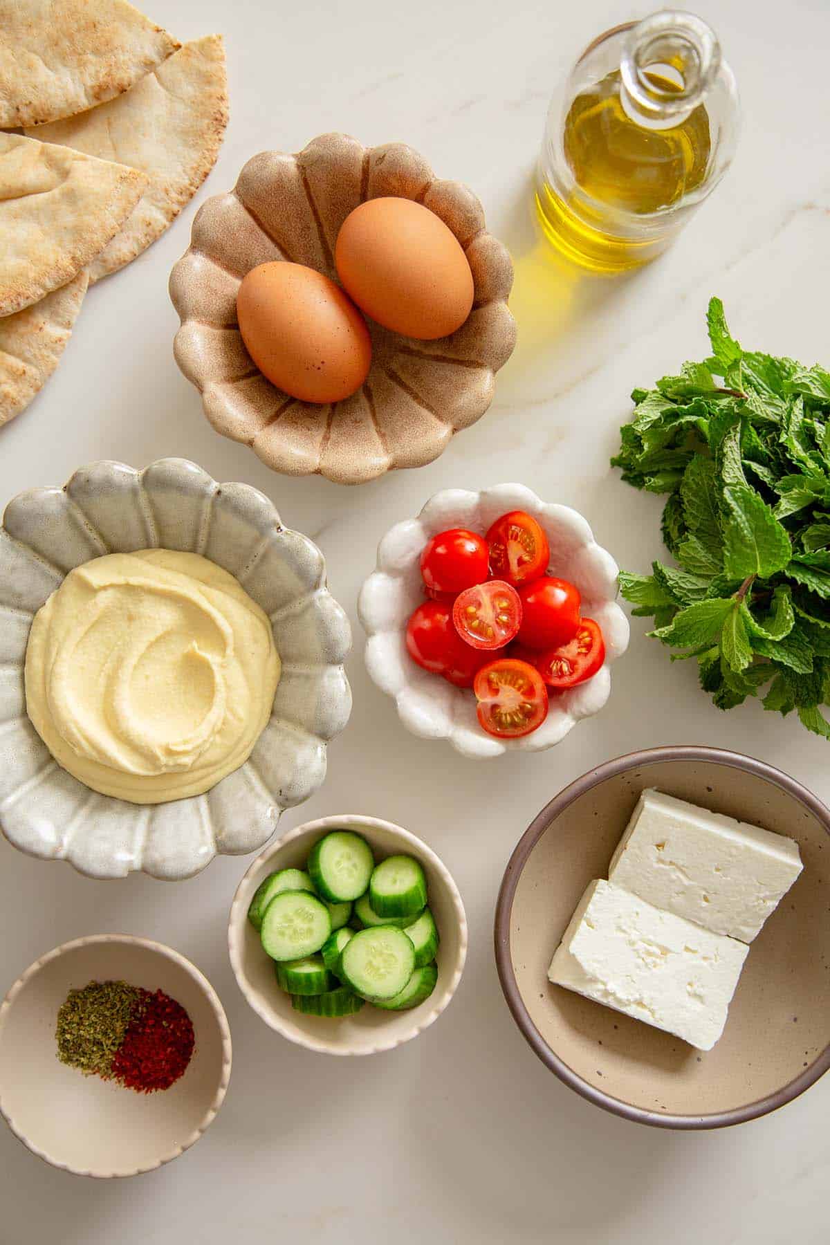 Overhead shot of the ingredients for a Mediterranean Breakfast Bowl, including hummus, eggs, tomatoes, cucumbers, feta, and spices.