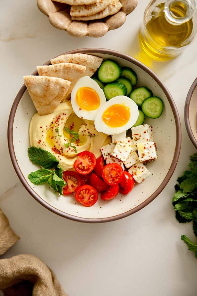 Top-down view of a Breakfast Bowl with hummus, eggs, feta, tomatoes, cucumbers, and pita bread.