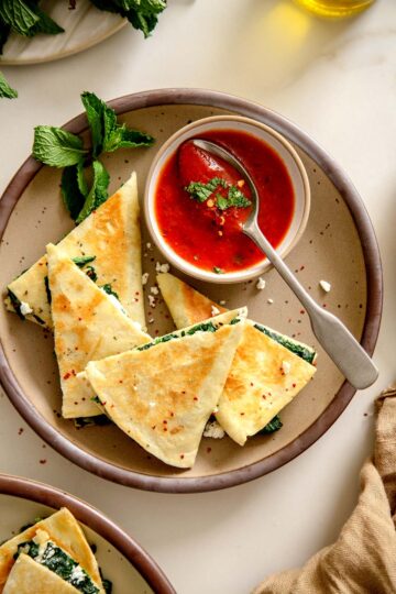 Close-up of golden brown Spanakopita Quesadilla wedges served with tomato sauce and fresh mint.