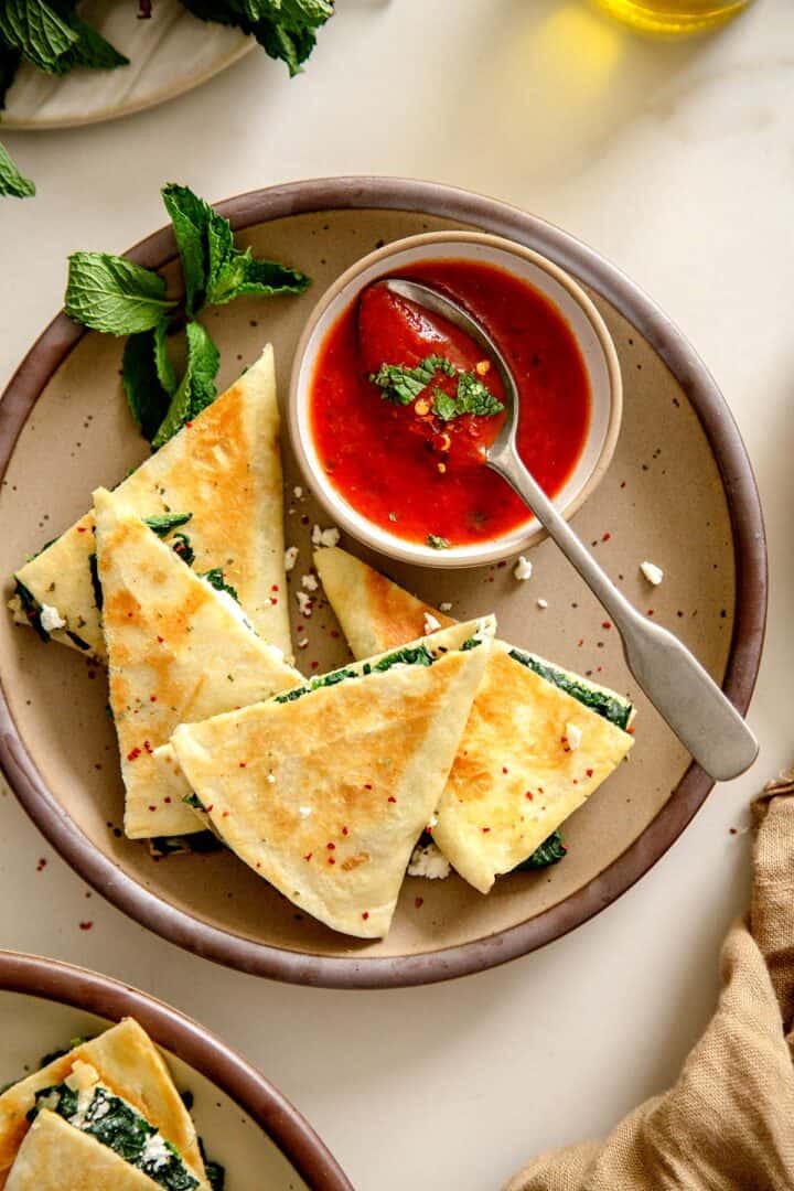 Close-up of golden brown Spanakopita Quesadilla wedges served with tomato sauce and fresh mint.