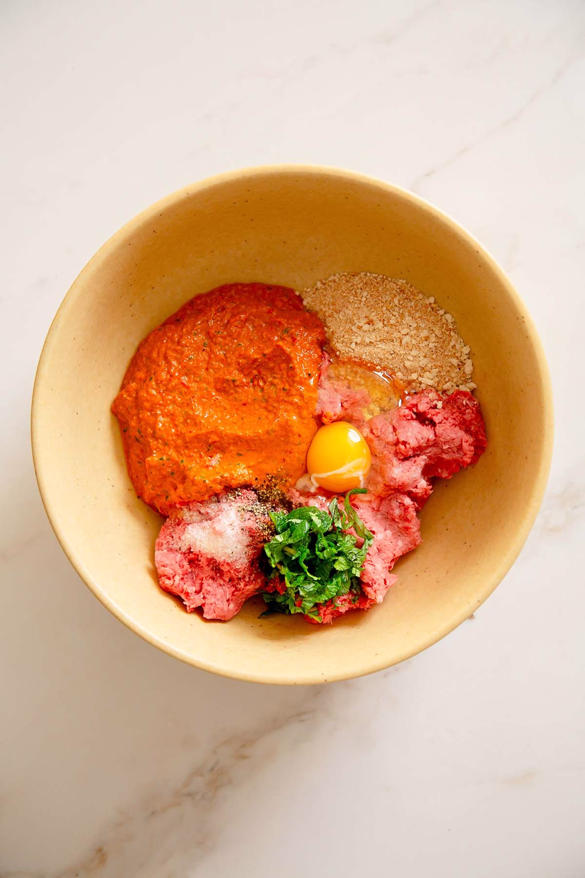  The bowl holds ground beef, a generous dollop of vibrant orange muhammara spread, breadcrumbs, fresh green herbs and a single egg yolk with its white. The ingredients are separated but ready for mixing.