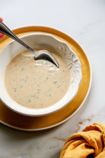 Bowl of zesty tahini sauce with garlic, fresh herbs, and za’atar, served with a spoon on a yellow plate