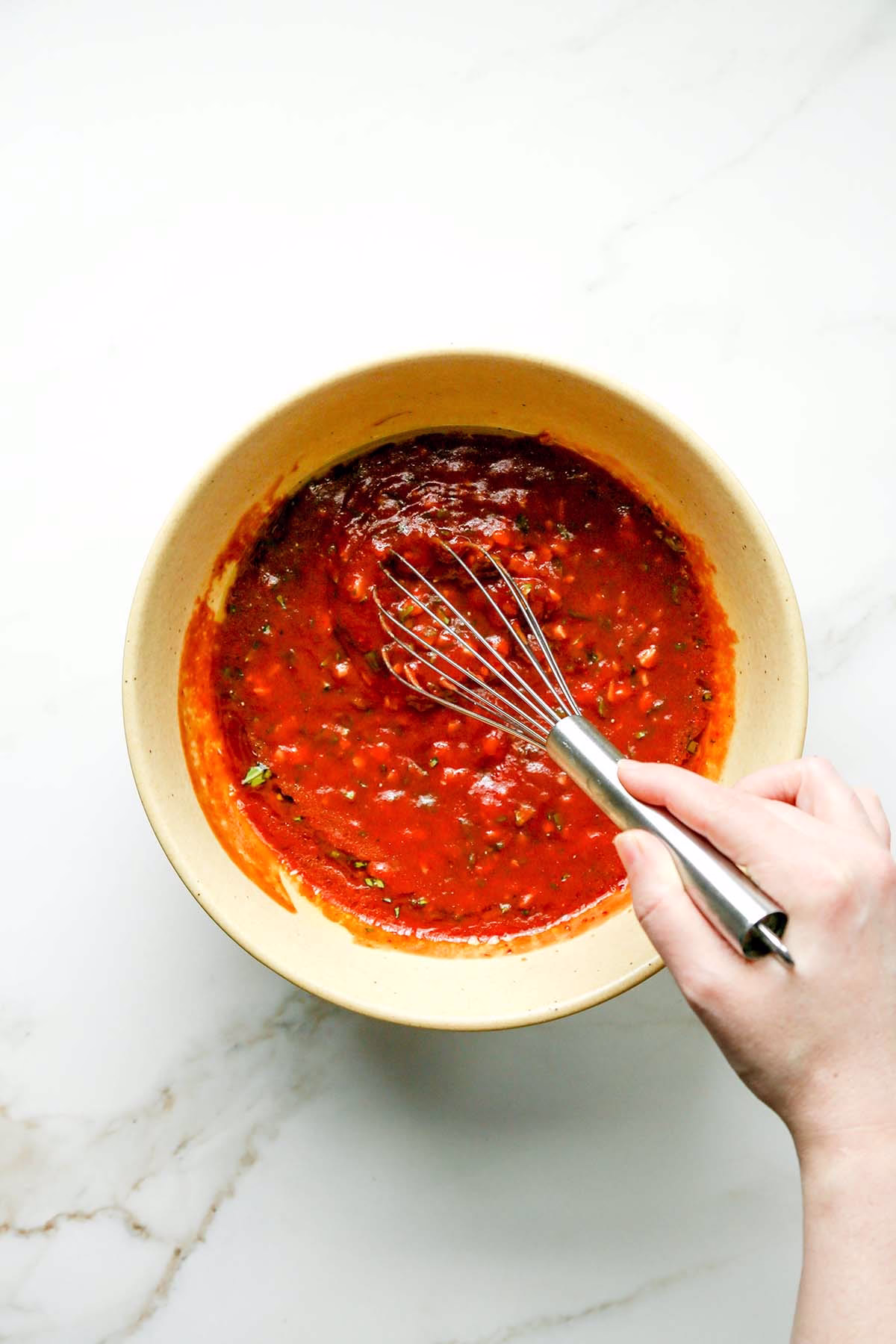 Hand whisking chopped garlic, basil, spices, and tomato sauce in a bowl for arrabbiata chicken marinade