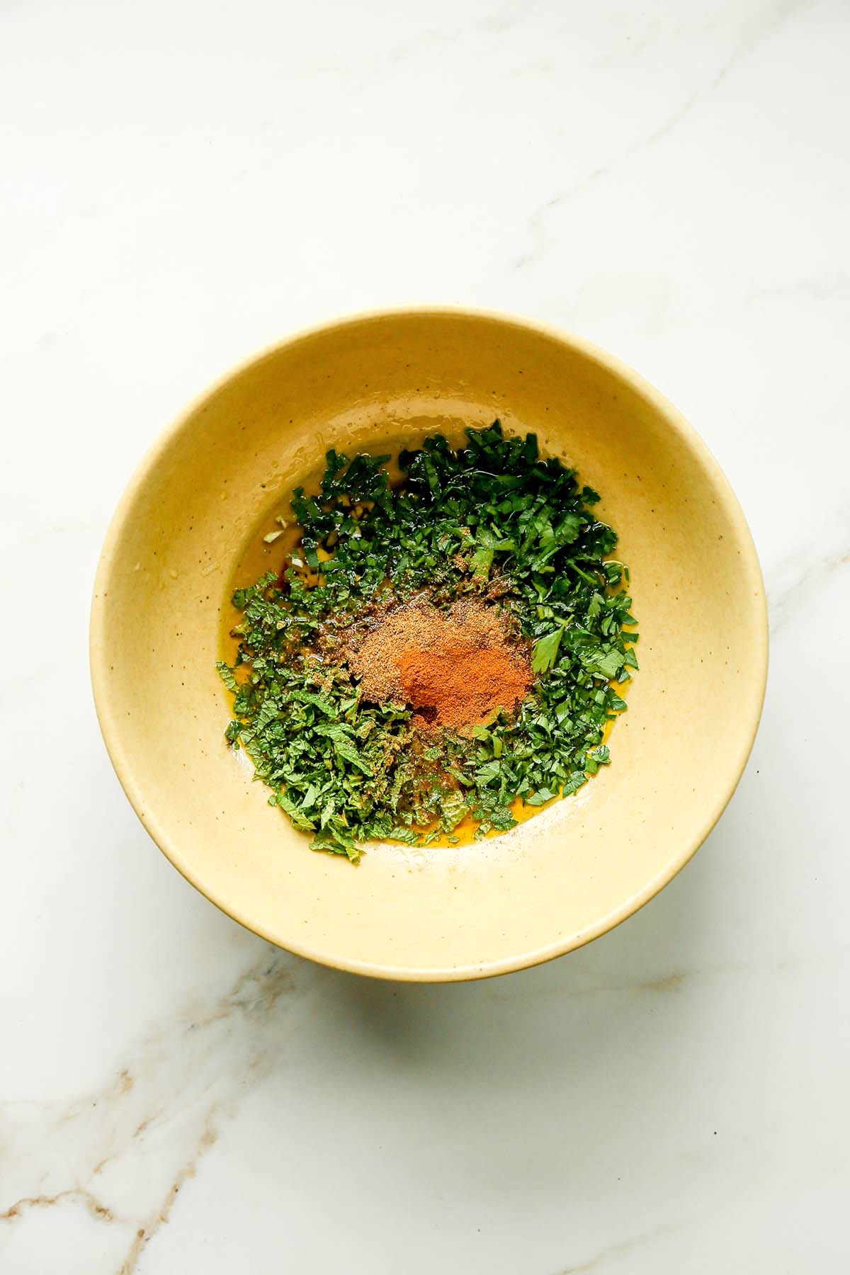 Unmixed Lebanese chicken marinade ingredients in a yellow bowl, including chopped parsley, mint, garlic, and shawarma spices.