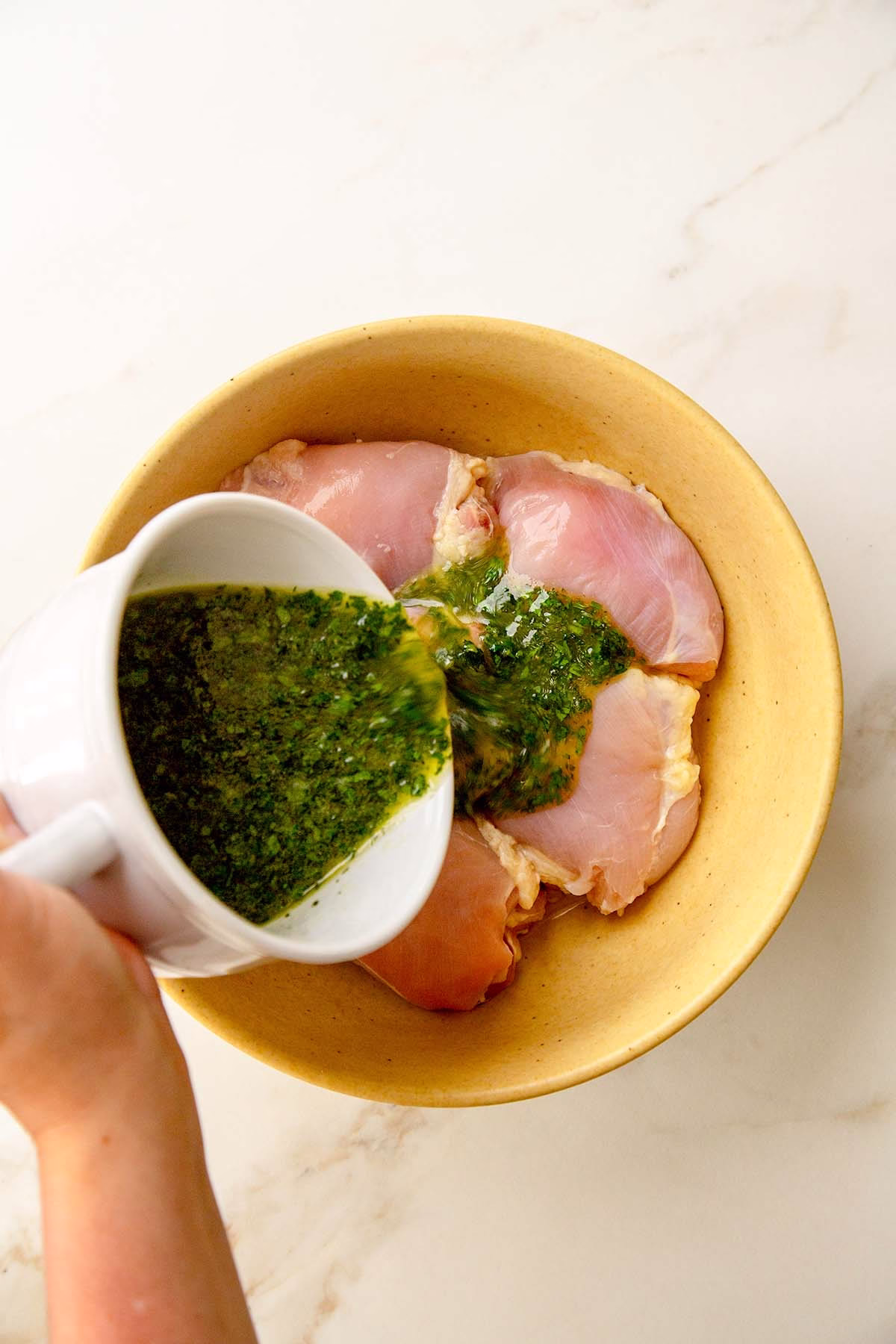 Bowl of lemony herb olive oil sauce poured over a yellow bowl of chicken thighs