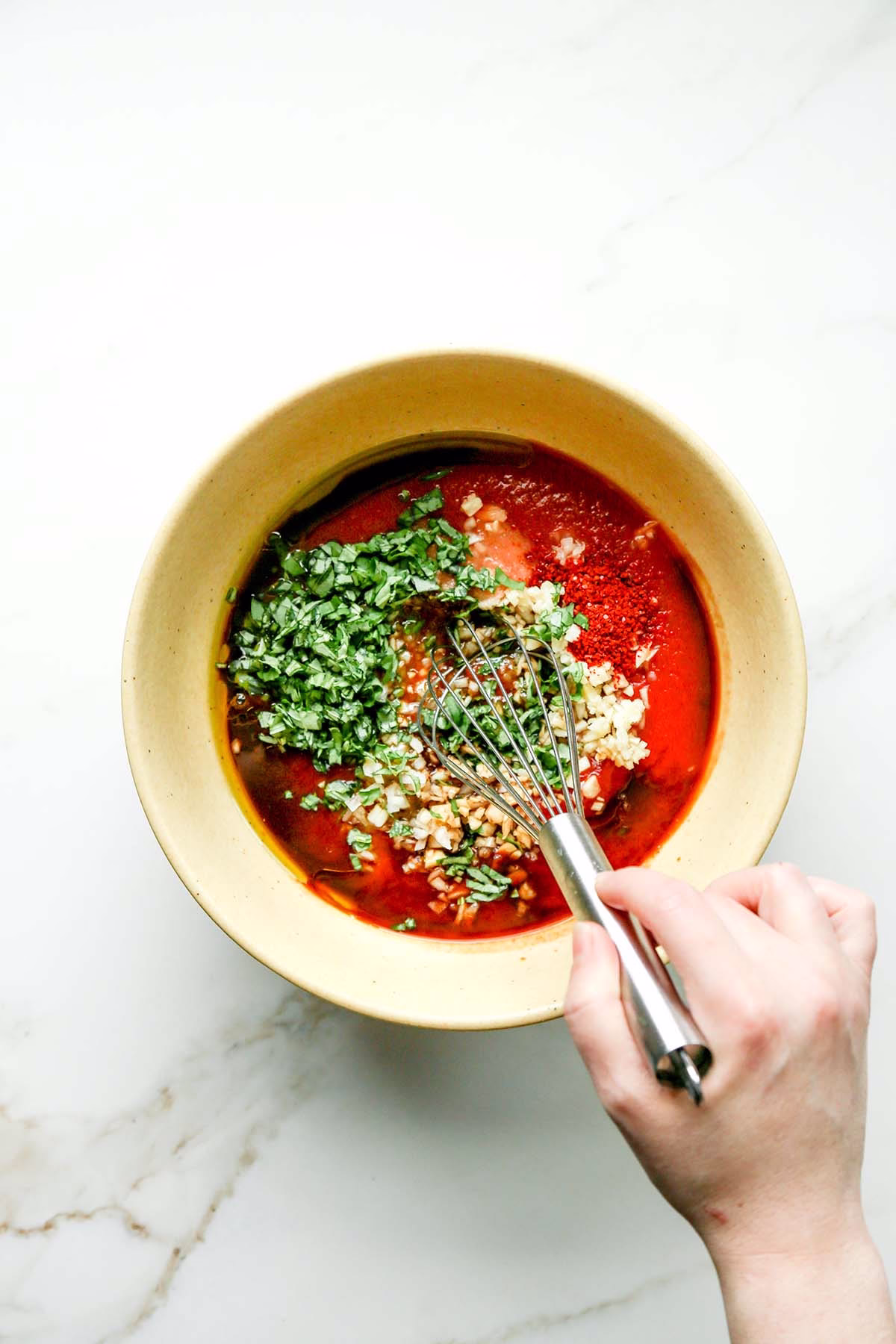 Hand whisking chopped garlic, basil, spices, and tomato sauce in a bowl for arrabbiata chicken marinade