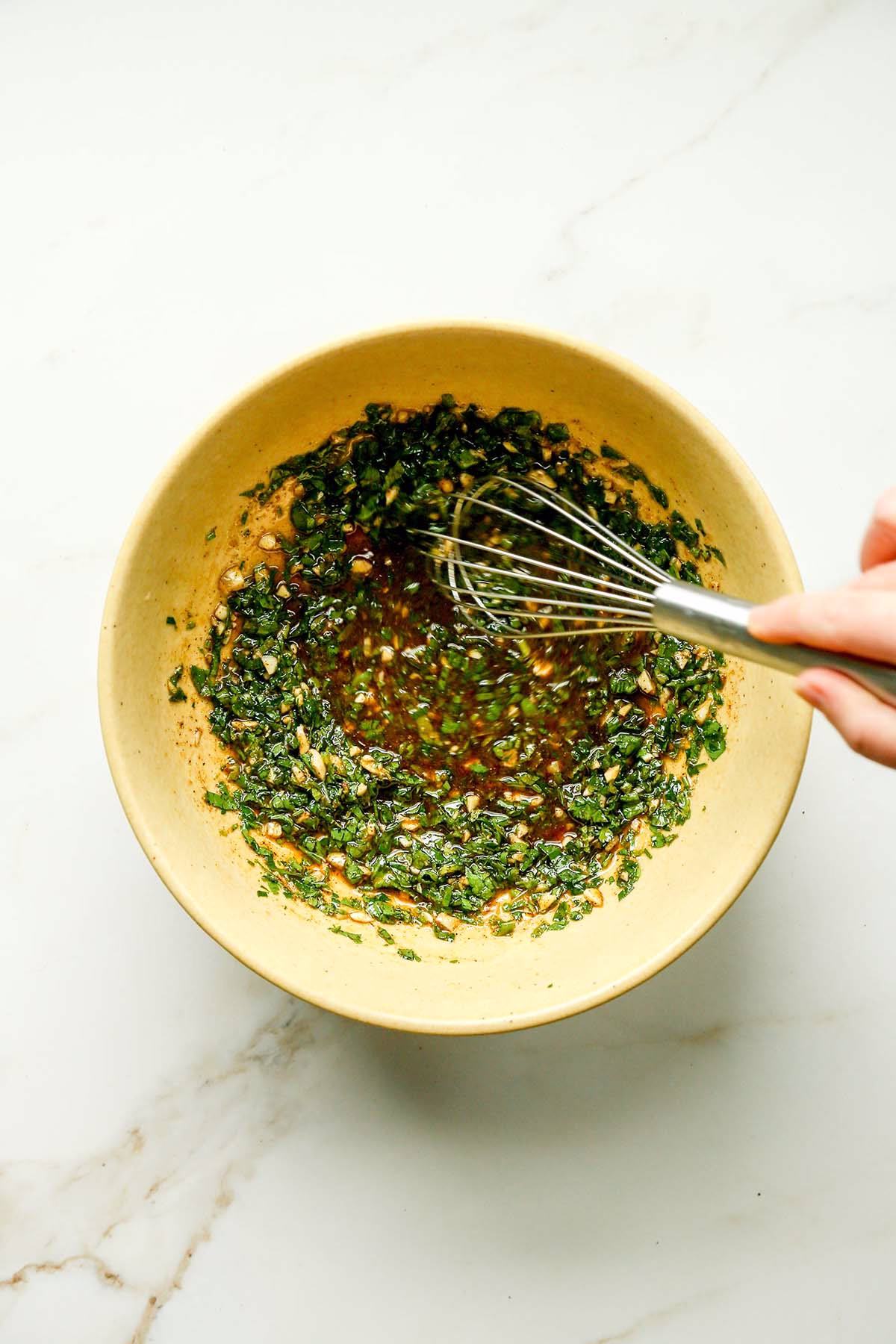 Hand whisking Lebanese chicken marinade with fresh herbs, garlic, olive oil, and pomegranate molasses in a yellow bowl.