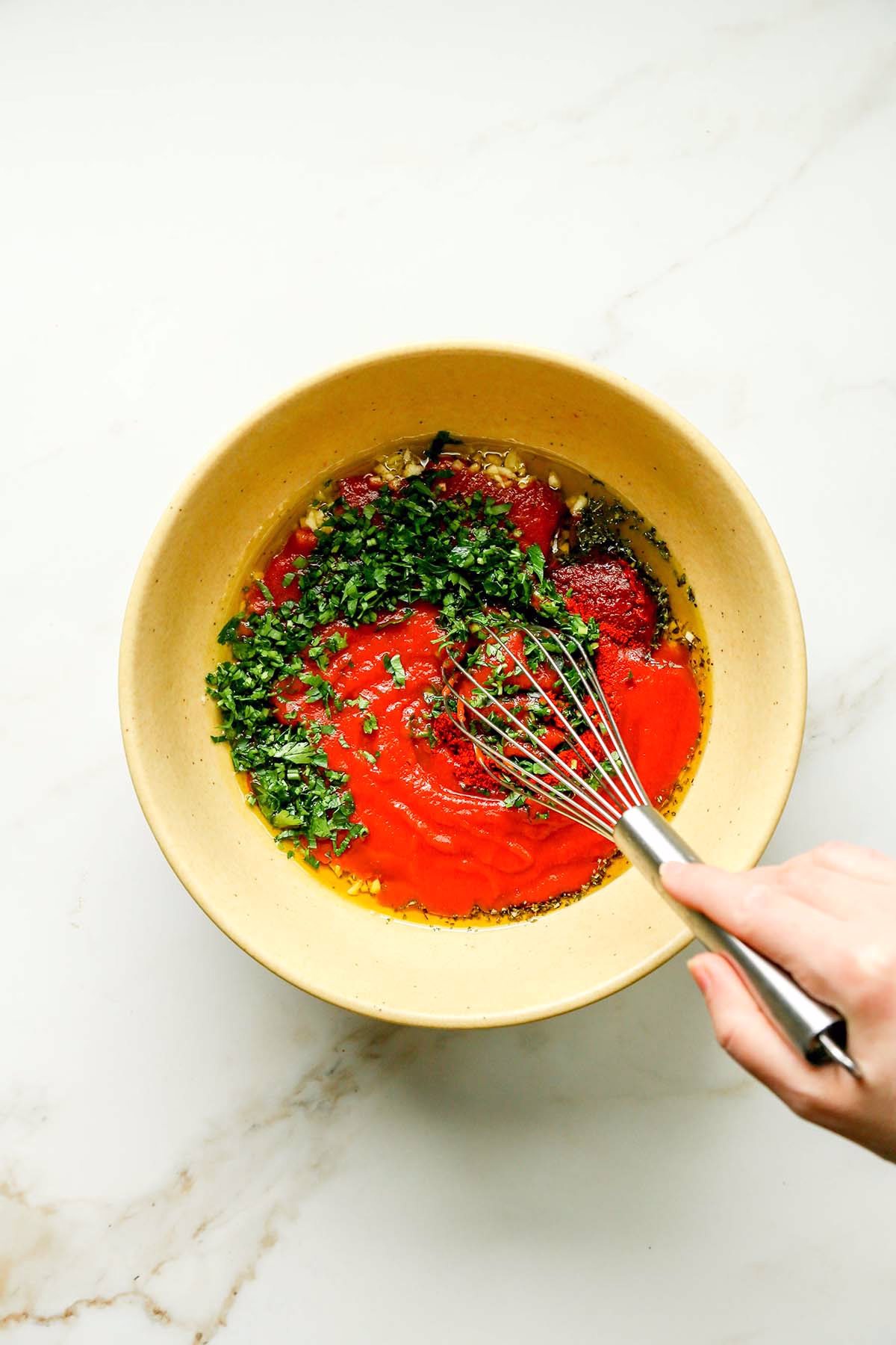 A hand whisking together tomato sauce, olive oil, and spices in a bowl.