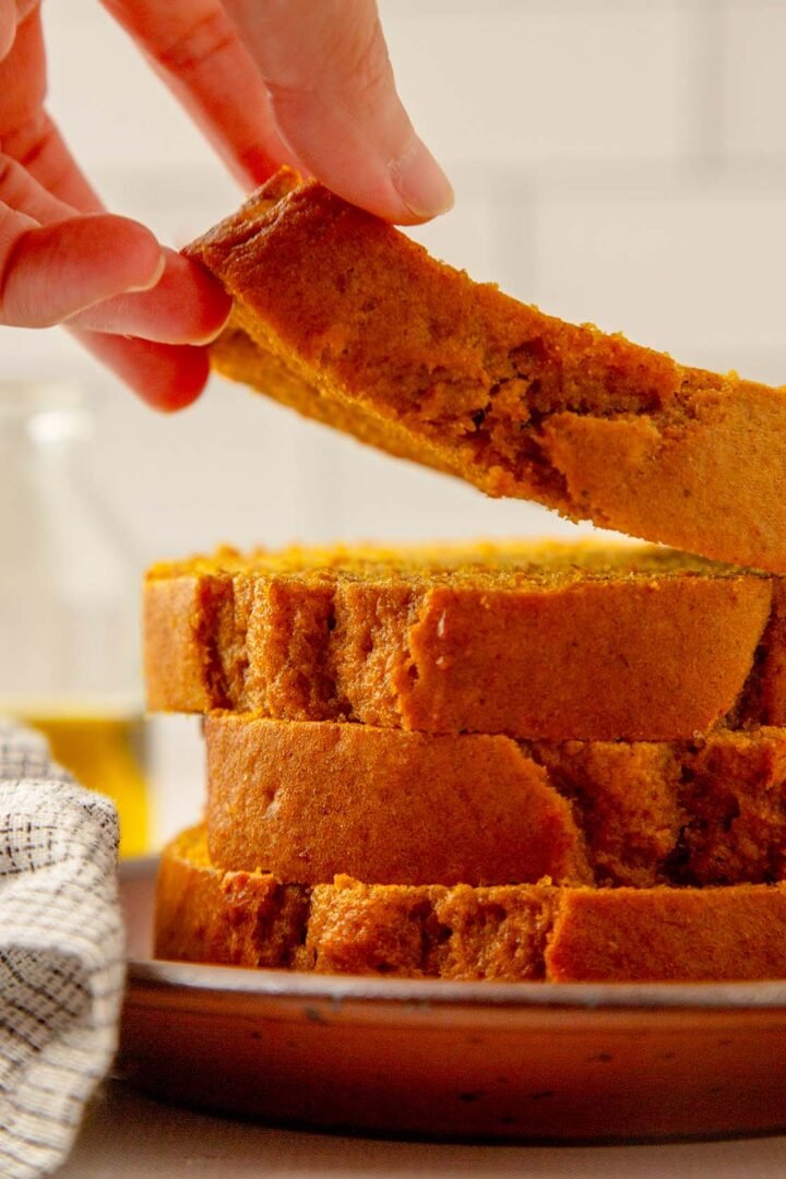 A hand lifts a slice of pumpkin bread from a stack of slices on a plate, highlighting the rich color and texture.