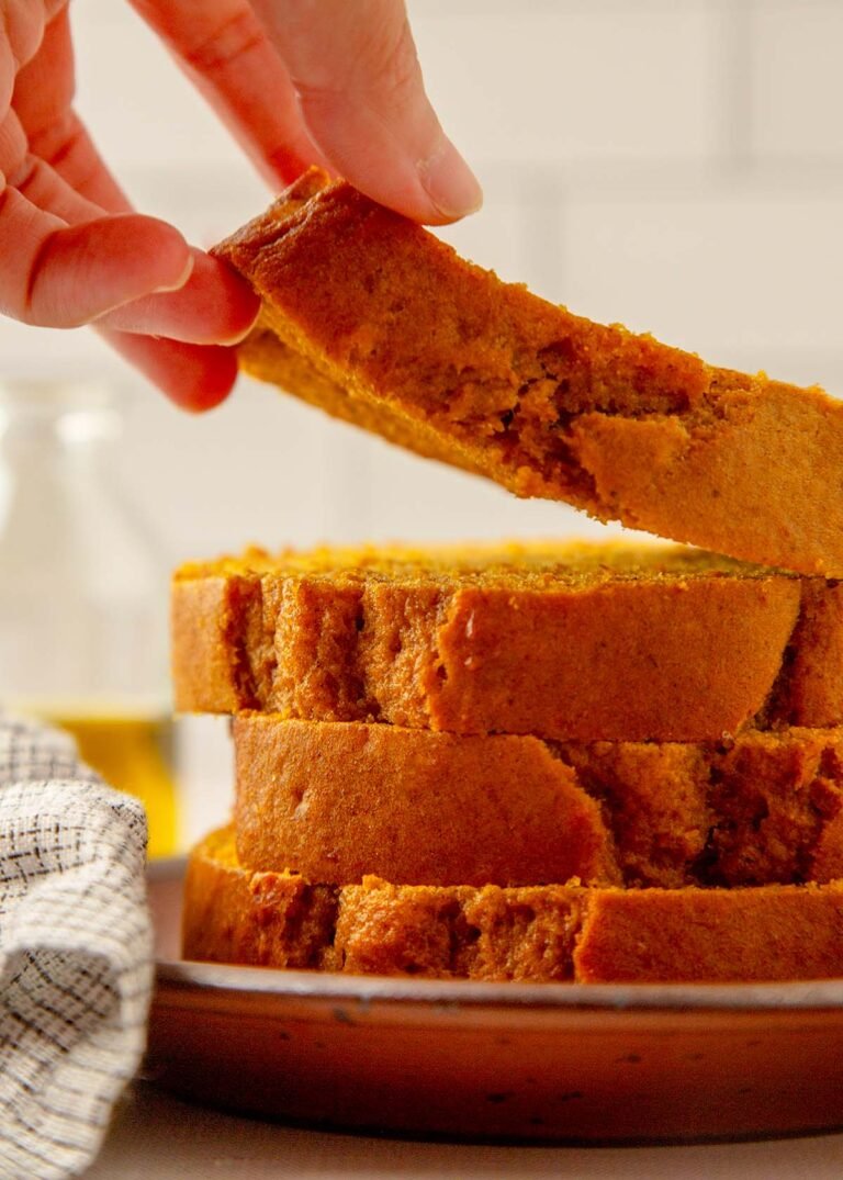 A hand lifts a slice of pumpkin bread from a stack of slices on a plate, highlighting the rich color and texture.