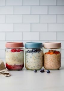 Glass jars filled with creamy overnight oats layered with fruit and toppings, showing Lemon & Blueberry, Raspberry Chia Jam, and Carrot Cake variations, ready for meal prep.