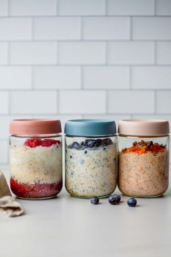 Glass jars filled with creamy overnight oats layered with fruit and toppings, showing Lemon & Blueberry, Raspberry Chia Jam, and Carrot Cake variations, ready for meal prep.