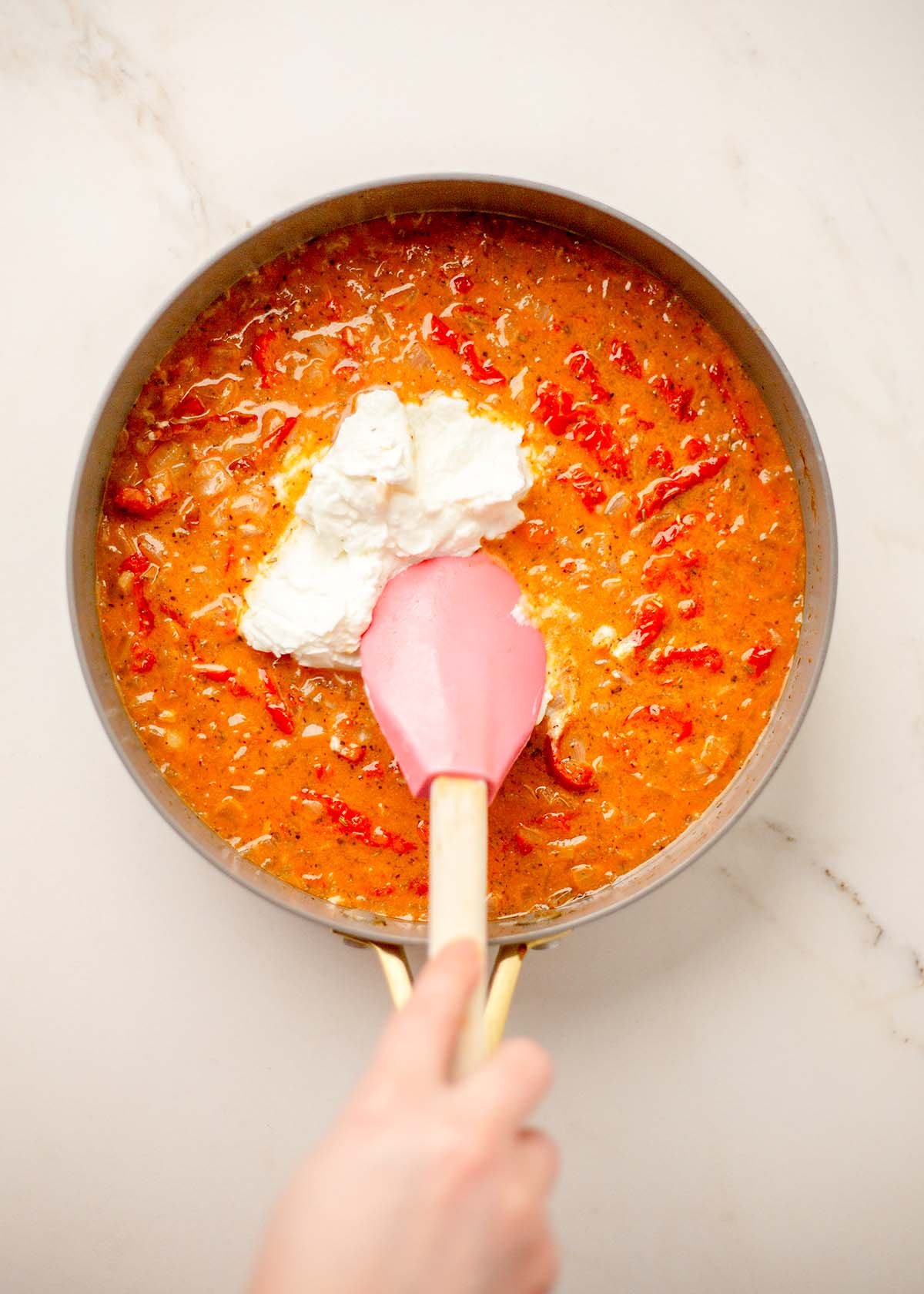 Person stirring creamy tomato sauce with Greek yogurt in a pan using a pink spatula.