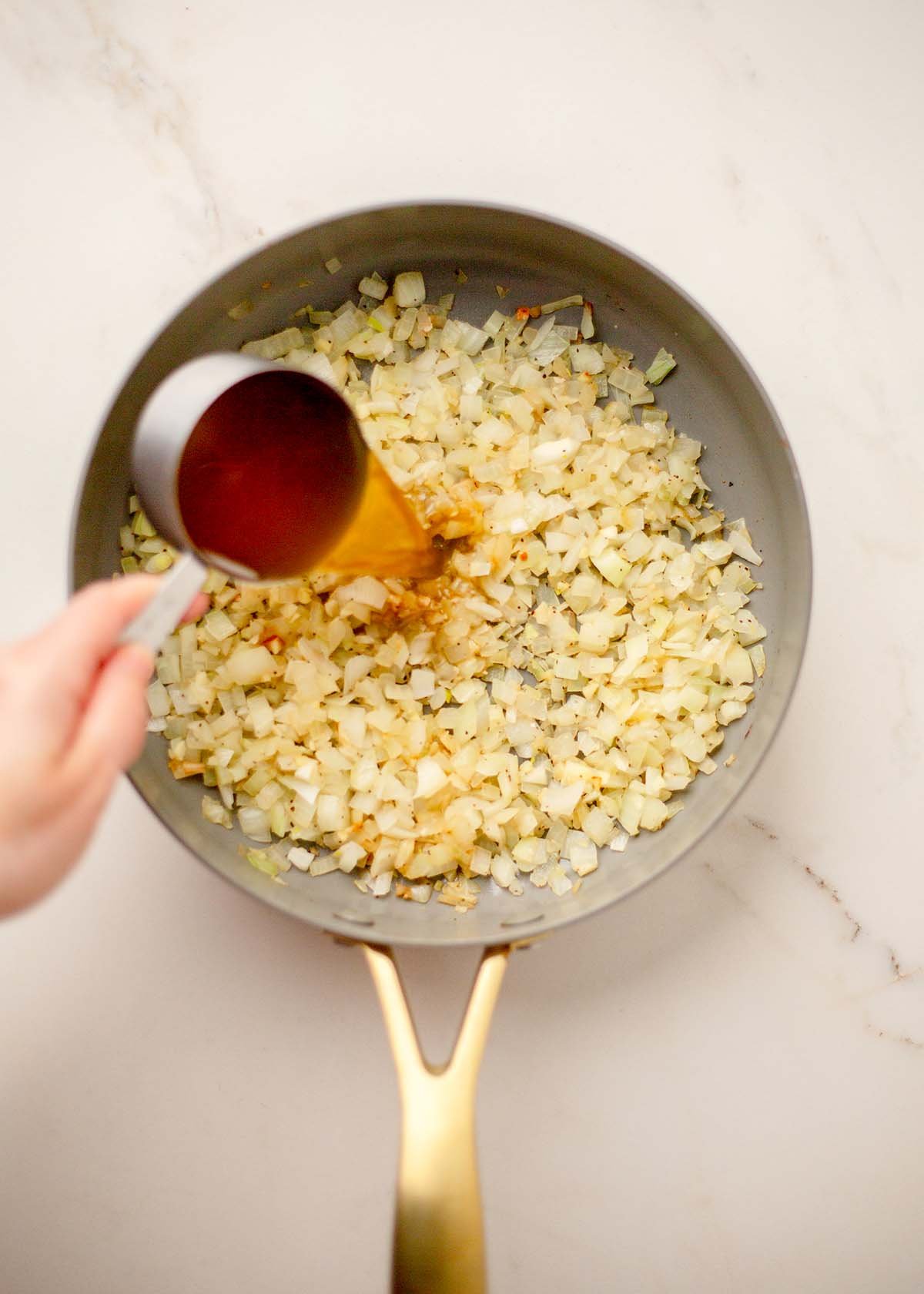A hand pours broth from a measuring cup into a skillet with sautéing chopped onions.
