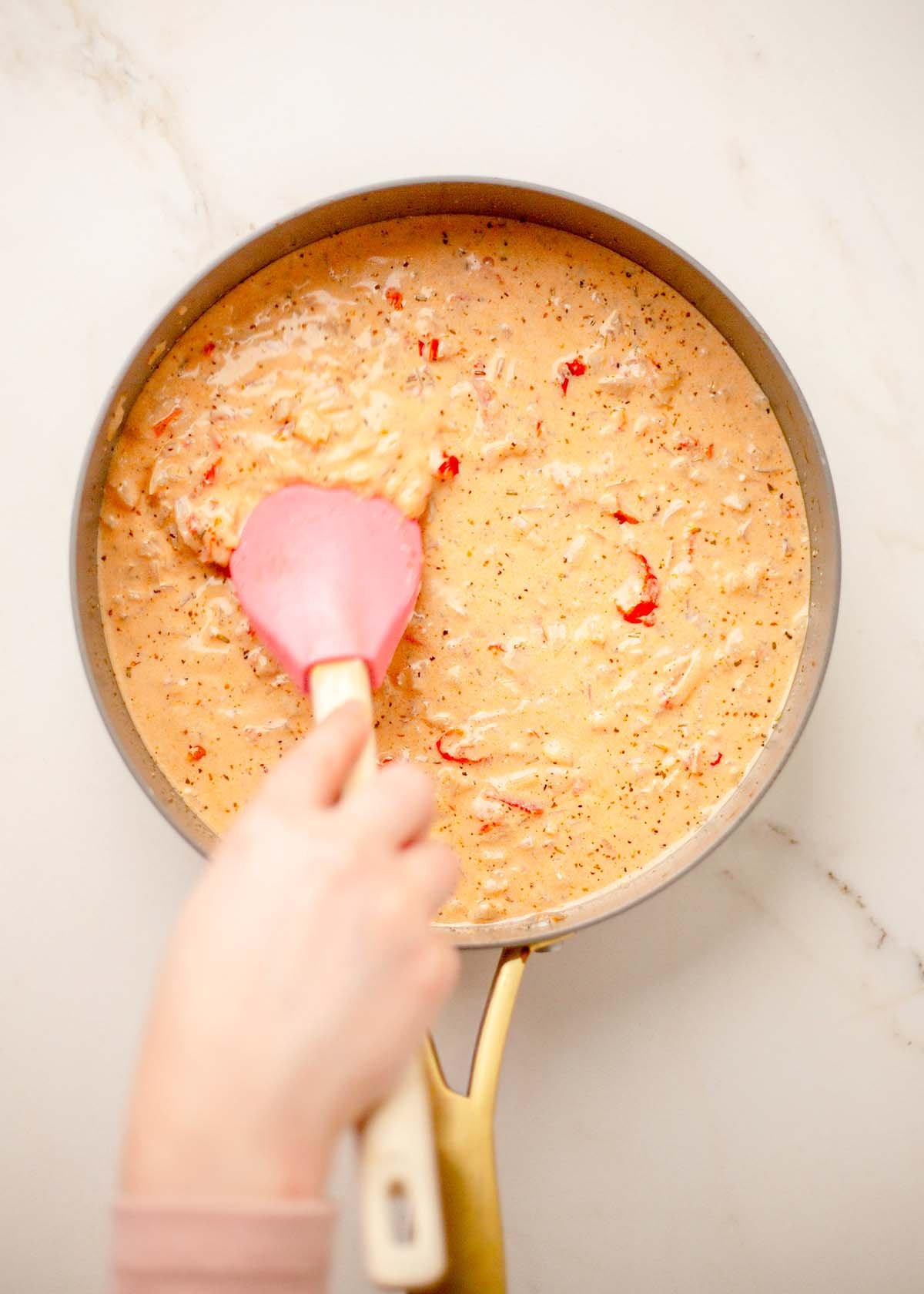 Person stirring creamy Mary Me sauce in a sauté pan using a pink spatula. The sauce is thick and speckled with herbs.